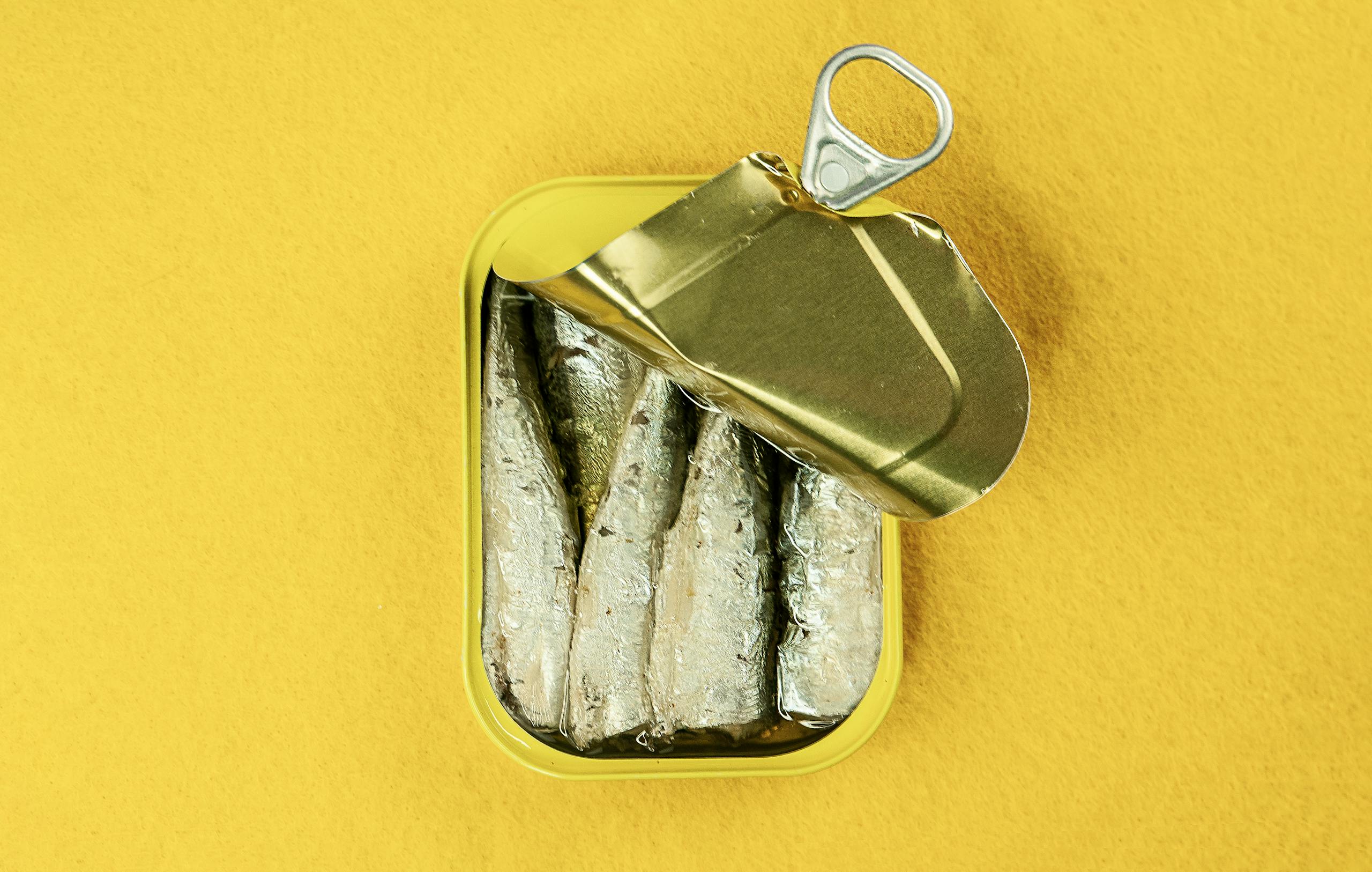 Metal can of sardines opened, revealing fish against a bright yellow background.